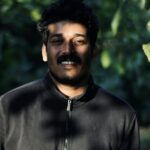 South Asian young man smiling in natural light outdoors, wearing a black jacket.